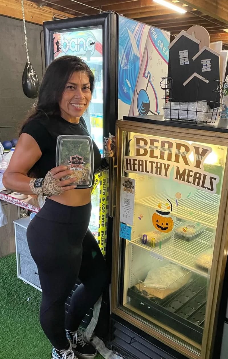 Beatriz holding a labeled meal container beside her branded Beary Healthy Meals fridge in a gym